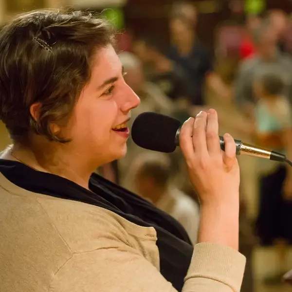 Rachel Shapiro Wallace, smiling into a microphone, with dancing people behind her.