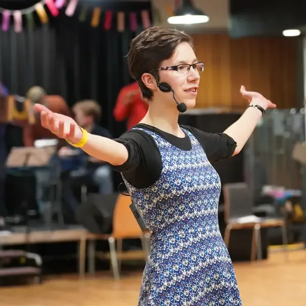 Lisa, a white woman in her thirties with short brown hair and glasses, is wearing a headset microphone and standing with her arms outstretched. Musicians are behind her on a stage (blurred).